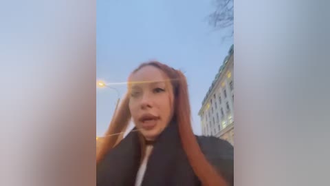 Media: Video of a young woman with long, reddish-brown hair, captured through a semi-transparent glass surface. She wears a black jacket. Background features a historic building and a cloudy sky.