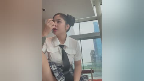 Media: Video of a young Asian woman with dark hair in a bun, wearing a white blouse, black tie, and plaid skirt, looking confused in a modern room with large windows and a city skyline outside.