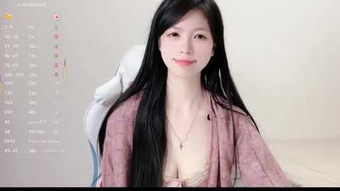 Media: Video of a fair-skinned, young East Asian woman with long black hair, wearing a pink robe over a beige lace bra, smiling softly. Background is a light-colored, plain room.