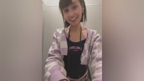 Media: Video of a young woman with straight, dark hair and a medium build, smiling. She wears a black tank top with \"CHILL\" in pink, and a purple and white striped hoodie. Background is plain white walls, creating a minimalistic setting.