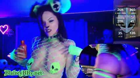 Media: Video of a woman in a green, fishnet, skull-print bodysuit with a black choker, wearing a blue panda hat, holding a red bottle, in a dimly lit club with a digital scoreboard in the background.