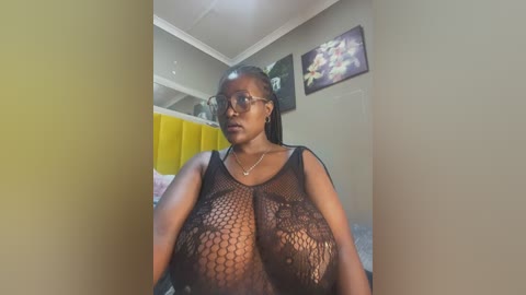 Media: Video of a dark-skinned woman with a large bust wearing a black fishnet top, standing in a bedroom with yellow walls and floral artwork.