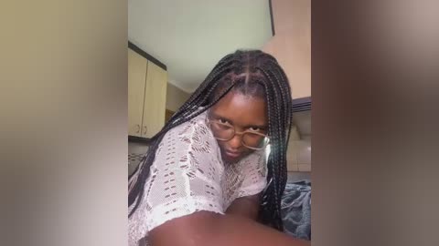 Media: Video of a Black woman with braided hair, wearing glasses and a white crochet top, leaning forward in a modern kitchen with beige cabinets and a gray countertop.