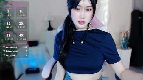 Media: Video of an Asian woman with long black hair in a ponytail, wearing a navy blue crop top, sitting at a gaming desk. Background shows a white desk, potted plants, and gaming peripherals.