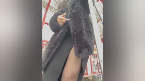 Media: Video of a person in a black coat with a high slit, revealing their thigh, standing in front of a red and white metal structure, likely a bus stop.