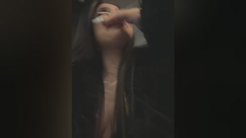 Media: A blurred video of a woman with long, dark hair, wearing a black jacket, covering her mouth with her hand, set in a dimly lit, possibly urban, background.