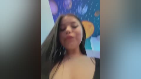 Media: Video of a young woman with long, wet hair, wearing a low-cut black top, standing in a shower with rain-like water droplets. Background features a colorful, abstract wall mural.