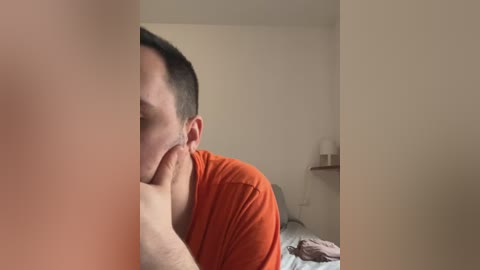 Media: A video shows a man with short dark hair, wearing an orange shirt, lying face down on a bed, with a blurred woman's hand touching his head. The background features a beige wall and a small nightstand.