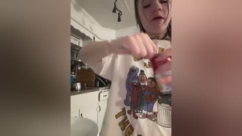 Media: Video of a woman with dark hair, wearing a white T-shirt featuring a cartoon family, preparing food in a kitchen with cabinets and a sink in the background.