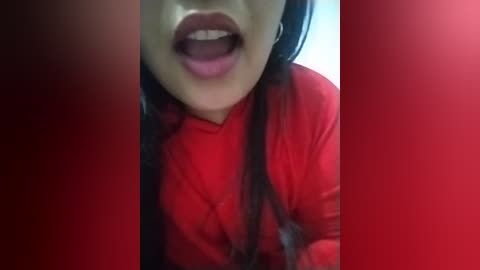 Media: A close-up video of a woman with light skin, long black hair, and full lips, wearing a bright red shirt, with a blurry red background. Her expression is open-mouthed, suggesting either laughter or surprise.