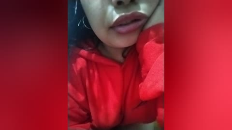 Media: Video of a woman with light brown skin, wearing a red hoodie, resting her head on her hand. She has full lips and is partially cropped, with a red background.