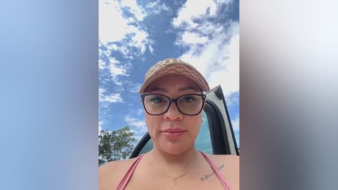 Media: Video of a young woman with fair skin, wearing black-rimmed glasses, a beige cap, and a pink bikini top, standing outdoors with a bright blue sky and scattered clouds in the background.