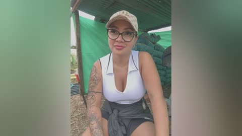 Media: Video of a Latina woman with medium skin tone, glasses, and a white polo shirt tied at the waist, sitting under a green tarp. She has a tattoo on her left arm.