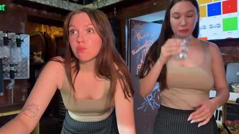 Media: Video of two women in a dimly lit bar. One woman, with a light complexion, long brown hair, and a tattoo on her left arm, is drinking from a bottle. The other woman, with a light complexion, long brown hair, and wearing a beige top, is standing next to her.