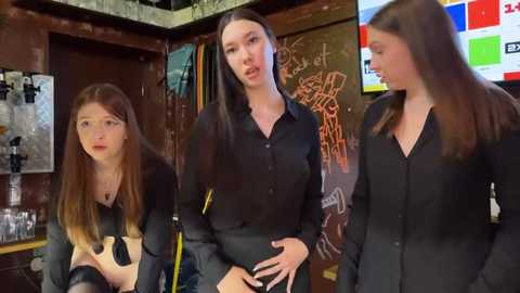 Media: Video of three women in black shirts, one with a yellow stripe, in a rustic, graffiti-covered room with a bar, shelves, and colorful posters.