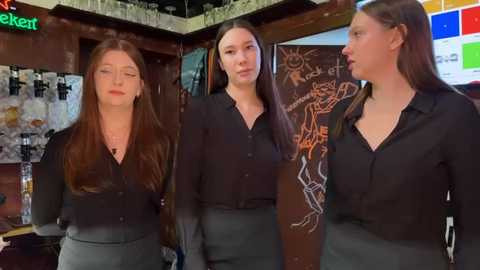 Media: Video of three young women with fair skin and long brown hair, dressed in black shirts and dark pants, standing in a cozy, dimly-lit bar with chalkboard wall art and colorful bar signs.