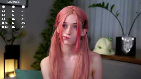 Media: A video of a young, fair-skinned woman with long, pink hair, wearing a black top, standing indoors with plants in the background.