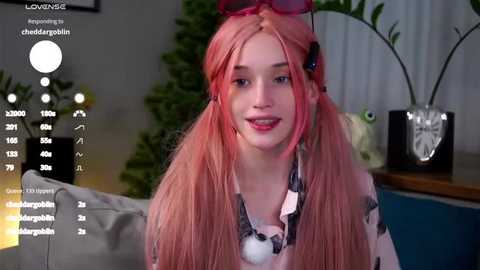 Media: Video of a young, fair-skinned woman with long pink hair in pigtails, wearing a black and white polka dot shirt, sitting indoors.