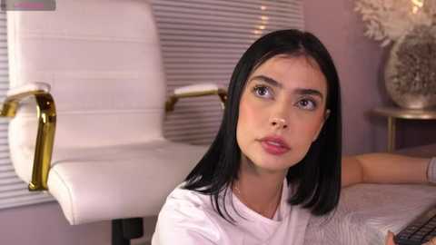 Media: Video of a young woman with straight black hair, light skin, and light makeup, sitting at a desk in a modern office with white and gold chairs and a lavender wall.