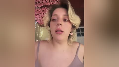 Media: Video of a fair-skinned, blonde woman with short, wavy hair, wearing a light purple spaghetti-strap top, standing in front of a backdrop of pink cherry blossom branches and fairy lights.