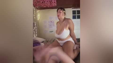 Media: Video of a curvy, light-skinned woman with short brown hair in white lingerie, kneeling on a bed in a cozy bedroom with pink fairy lights, calendars, and a window.