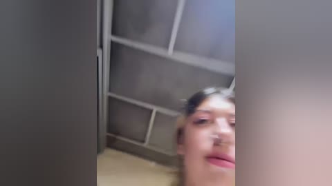 Media: Video of a young woman with fair skin, brown hair, and light makeup, smiling and sticking out her tongue, captured from a low angle in a small, dimly lit room with metallic paneling.