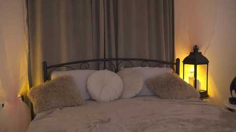 Media: A cozy, dimly-lit bedroom video features a black wrought iron bed with white pillows and a beige throw, flanked by two side lamps with soft yellow light, and grey curtains in the background.