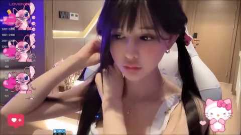 Media: A video of a young Asian woman with long black hair styled in pigtails, wearing a low-cut white top, captured in a cozy, well-lit room with a pink Hello Kitty decoration.