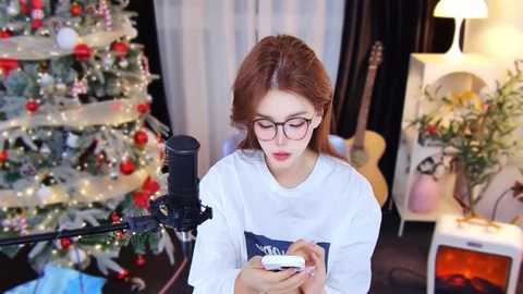 Media: Video of a young woman with red hair, wearing glasses and a white sweater, recording a song with a microphone on a black stand, in a cozy room with a decorated Christmas tree, guitar, and potted plants.