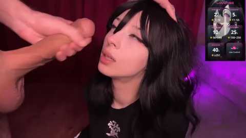 Media: Video of a young Asian woman with long black hair, wearing a black shirt, kneeling with a man's erect penis in her hand, in a dimly lit room with a digital screen displaying a video game interface.