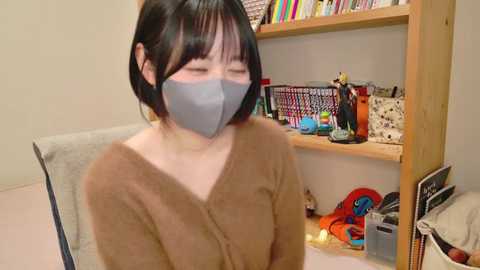 Media: Video of an Asian woman with short black hair, wearing a gray face mask, brown sweater, and sitting in a cluttered, bookshelf-filled room.