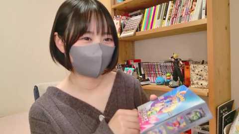 Media: Video of an Asian woman with short black hair, wearing a gray face mask, gray cardigan, and holding a colorful, cartoon-themed gift box in a bookshelf-filled room.