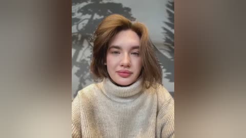 Media: Video of a young woman with shoulder-length, tousled brown hair, wearing a beige, ribbed turtleneck sweater, against a blurred, grayscale background resembling a tree.