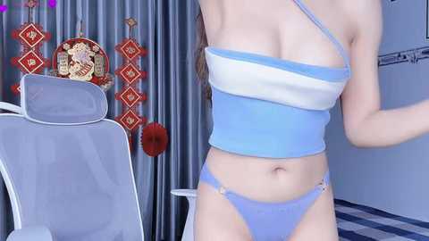 Media: Video of a fair-skinned woman with large breasts, wearing a light blue, one-shoulder crop top and matching thong, standing in a room with grey curtains and decorative hanging red and gold ornaments.