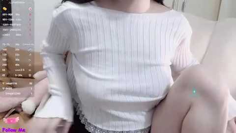 Media: Video of a woman with fair skin, wearing a white ribbed long-sleeve top and grey lace panties. She sits on a white couch, her left leg bent, with a blue-green light illuminating her thigh.