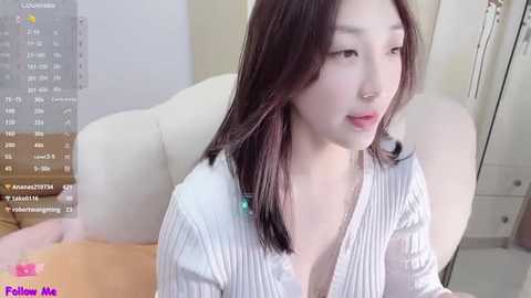 Media: Video of a young East Asian woman with long, dark hair, wearing a white, ribbed cardigan, sitting on a beige couch, with a digital clock and \"Follow Me\" text in the background.