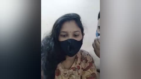 Media: Video of a South Asian woman with long black hair, wearing a floral blouse and black face mask, capturing her reflection in a mirror, surrounded by vertical white and black bars.