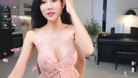 Media: A video of a young East Asian woman with long black hair, fair skin, and wearing a sheer, pink lace lingerie top, standing in a modern living room with a piano and plants.