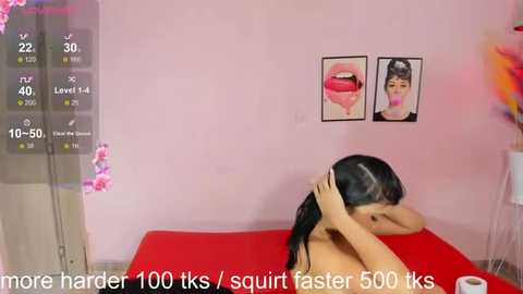 Media: A video of a young Asian woman with long black hair, topless, lying face-down on a red bed, in a pink room with a digital readout.