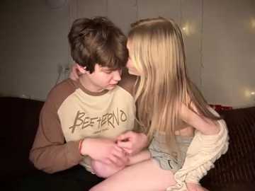 Media: Video of a young boy and girl sitting close on a couch, sharing a tender moment. He wears a beige and brown shirt, while she's in a white dress. Background is dimly lit.