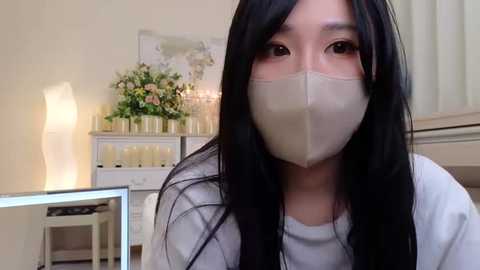 Media: Video of an Asian woman with long black hair and light skin, wearing a beige face mask and white shirt, in a softly lit room with floral arrangements and candles on a white dresser.