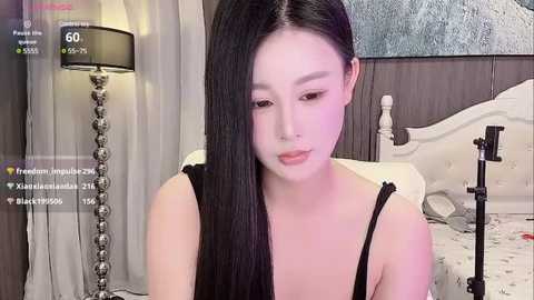 Media: A video of a young Asian woman with long black hair, light skin, and full makeup, sitting on a bed, wearing a black top, in a bedroom with a wooden headboard, white bedding, and a lamp.