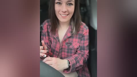 Media: Video of a smiling young woman with shoulder-length brown hair, wearing a red and black plaid shirt, adjusting a smartphone inside a car.