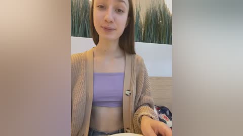 Media: Video of a young woman with straight, brown hair, wearing a light purple crop top and beige cardigan, seated indoors with a modern, minimalist background.