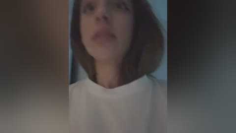 Media: A video of a young woman with light skin and shoulder-length brown hair, wearing a white shirt, taken from a low angle. The background is blurred, with a soft, muted color palette.