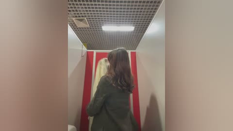 Media: Video of a narrow hallway with beige walls and a grid-patterned ceiling. A woman with shoulder-length brown hair, wearing a green coat, walks away from the camera. Red-striped door in the background.