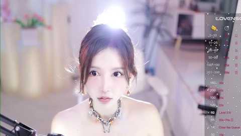 Media: Video of a young, fair-skinned woman with shoulder-length brown hair, wearing a silver necklace, captured from a selfie stick, in a softly lit room with blurred background.