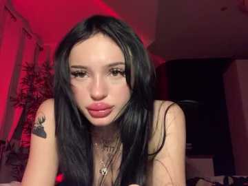 Video of a pale-skinned woman with long, straight black hair, wearing a black top, leaning forward, with a tattoo on her left shoulder. Background has red lighting, a potted plant, and a dark curtain.