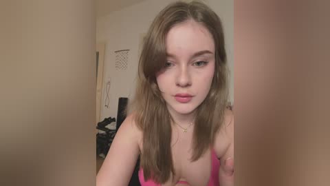 Media: A video of a young Caucasian woman with fair skin, brown hair, and green eyes, wearing a pink tank top, standing in a dimly lit hallway.