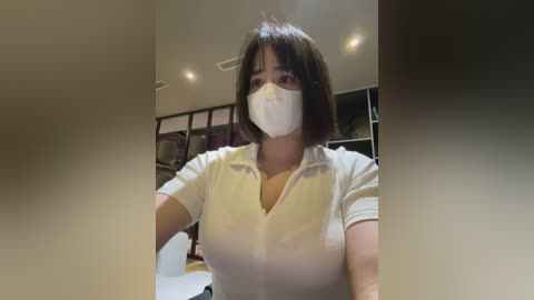 Media: A video of a woman with short dark hair, wearing a white mask and a white short-sleeved shirt, standing in a dimly lit, modern kitchen with recessed lights and dark cabinets.